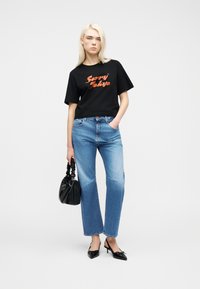 Ξανθιά γυναίκα όρθια, φοράει μαύρο μπλουζάκι με την επιγραφή "Sorry Tokyo", ανοιχτόχρωμο τζιν, μαύρες χαμηλές γόβες, κρατώντας μαύρη τσάντα με μαζεμένη λαβή.