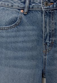 Gros plan sur un jean en denim bleu montrant une poche avant, une boucle de ceinture, des coutures, des rivets métalliques et les détails de la fermeture par bouton.