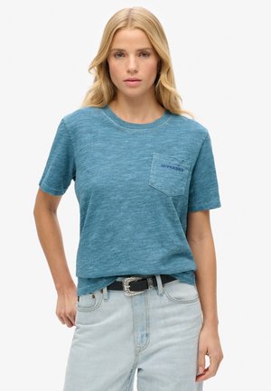 T-shirt turquoise à manches courtes en tissu texturé, avec une poche poitrine arborant le logo "SUPERDRY". Associé à un jean clair et une ceinture noire.