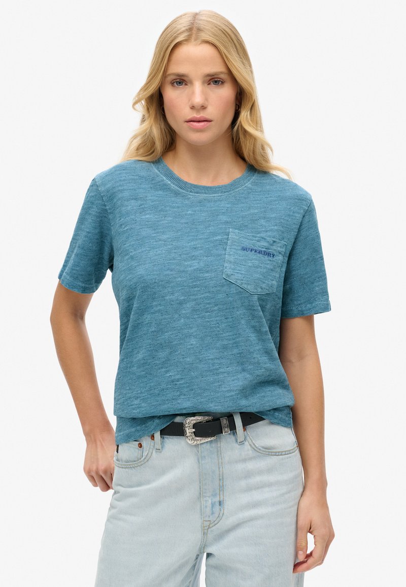 Tyrkis kortærmet T-shirt lavet af tekstureret stof, med en brystlomme med "SUPERDRY" logo. Kombineret med lyse jeans og et sort bælte.