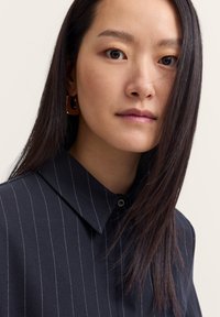 Collared shirt in dark fabric with white vertical stripes and a button at the collar; featuring long straight hair and gold hoop earrings.