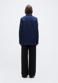 Άτομο που στέκεται με την πλάτη προς το μέρος μας, φοράει μακρυμάνικο πουκάμισο σκούρου μπλε χρώματος και μαύρο παντελόνι φαρδιάς γραμμής, με απλό λευκό φόντο.