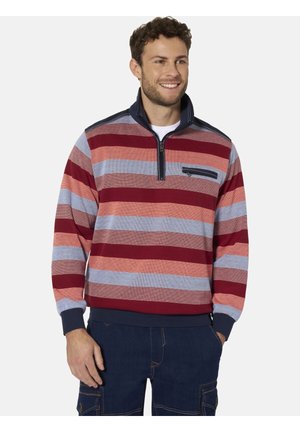 Mann lächelt, trägt einen langärmeligen Pullover mit Viertelreißverschluss mit horizontalen roten, orangefarbenen und blauen Streifen sowie dunkelblaue Hosen mit Taschen.