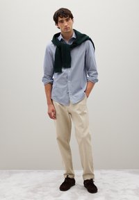 Camisa de botones a rayas azul y blanca, pantalones crema, zapatos marrón oscuro y un suéter verde oscuro atado alrededor de los hombros.