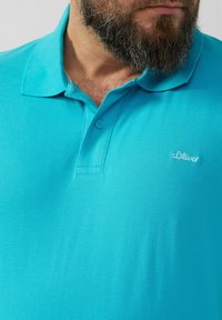 Türkise Polo-Shirt aus strukturiertem Stoff, mit Kragen, zwei Knöpfen und einem kleinen weißen s.Oliver-Logo auf der linken Seite.