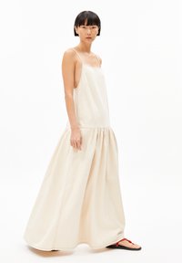 Beige Baumwoll-Maxikleid mit einem taillierten Oberteil, dünnen Trägern und einem gerafften, voluminösen Rock. Getragen mit schwarzen Sandalen mit roten Akzenten.