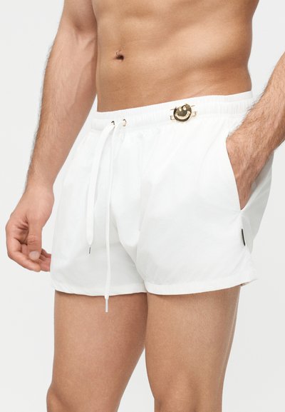 Άνδρας που φοράει λευκά μαγιό σορτς με ελαστική μέση, κορδόνι ρύθμισης, πλαϊνές τσέπες και ένα μικρό χρυσό καρφίτσα με χαμογελαστό πρόσωπο στη μέση.