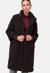 Manteau long et brun, duveteux, avec un col châle, pr�ésentant des poignets en côtes marron. Porté par-dessus un col roulé bordeaux, avec une coupe décontractée.