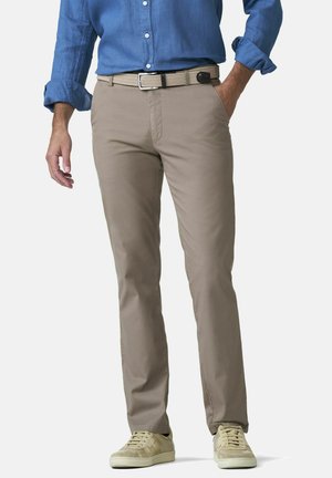 Man draagt beige broek, geweven beige riem, lichtbeige sneakers en een blauw opgerolde lange mouwen overhemd, staat met één hand in de zak.