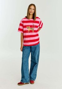 Oversized tričко s červenými a růžovými pruhy s grafikou kočky. Kombinováno s volnými modrými džíny a červenými botami.