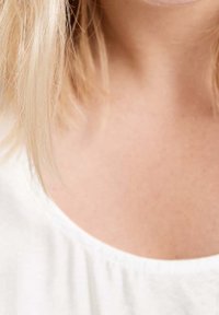 Des cheveux blonds tombent sur l'épaule d'une personne portant un haut blanc à col rond, laissant apparaître le cou et le haut de la poitrine.