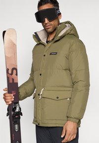 Muškarac u maslinasto zelenoj puf jakni i crnim skijaškim naočalama drži ružičaste i crne skije ispred jednostavne pozadine.