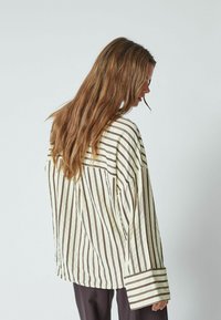 Chemise surdimensionnée rayée en jaune pâle avec des lignes verticales marron foncé, dotée de larges manches et d'une coupe décontractée. La texture semble lisse.
