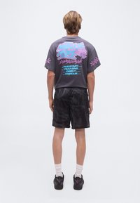 Νέος άνδρας που στέκεται με την πλάτη του ορατή, φορώντας σκούρο μπλουζάκι με πολύχρωμο κείμενο και γραφικά αστεριών, μαύρη σορτς με σχέδια, λευκές κάλτσες και μαύρα αθλητικά παπούτσια.