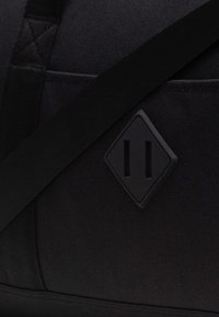 Sac de voyage en tissu noir avec une surface texturée, une sangle ajustable et un logo en forme de diamant avec deux fentes verticales.