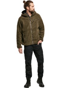 Veste à capuche verte olive doublée en polaire avec fermeture éclair sur le devant, poches latérales et tissu texturé, associée à un pantalon cargo noir et des bottes.
