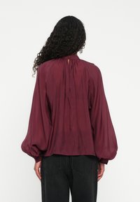 Personne aux longs cheveux bouclés portant un chemisier ample à manches longues bordeaux et un pantalon noir, vue de dos sur fond blanc.