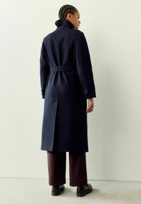 Manteau en mélange de laine bleu marine avec un col montant, une taille ceinturée et des fentes latérales. Porté sur un pantalon bordeaux et des chaussures noires à finition brillante.