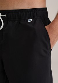Schwarze Badehosenshorts mit einem elastischen Bund, weißem Kordelzug, Seitentaschen und einem kleinen Palmenlogo auf der linken Hüfte.