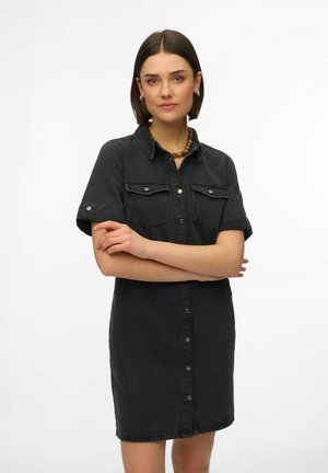 Rövid sötét hajú nő, fekete gombos farmerruhát visel, amit felhajtott ujjakkal, és fa gyöngy nyakláncot, karjait összefonva, semleges háttér előtt.