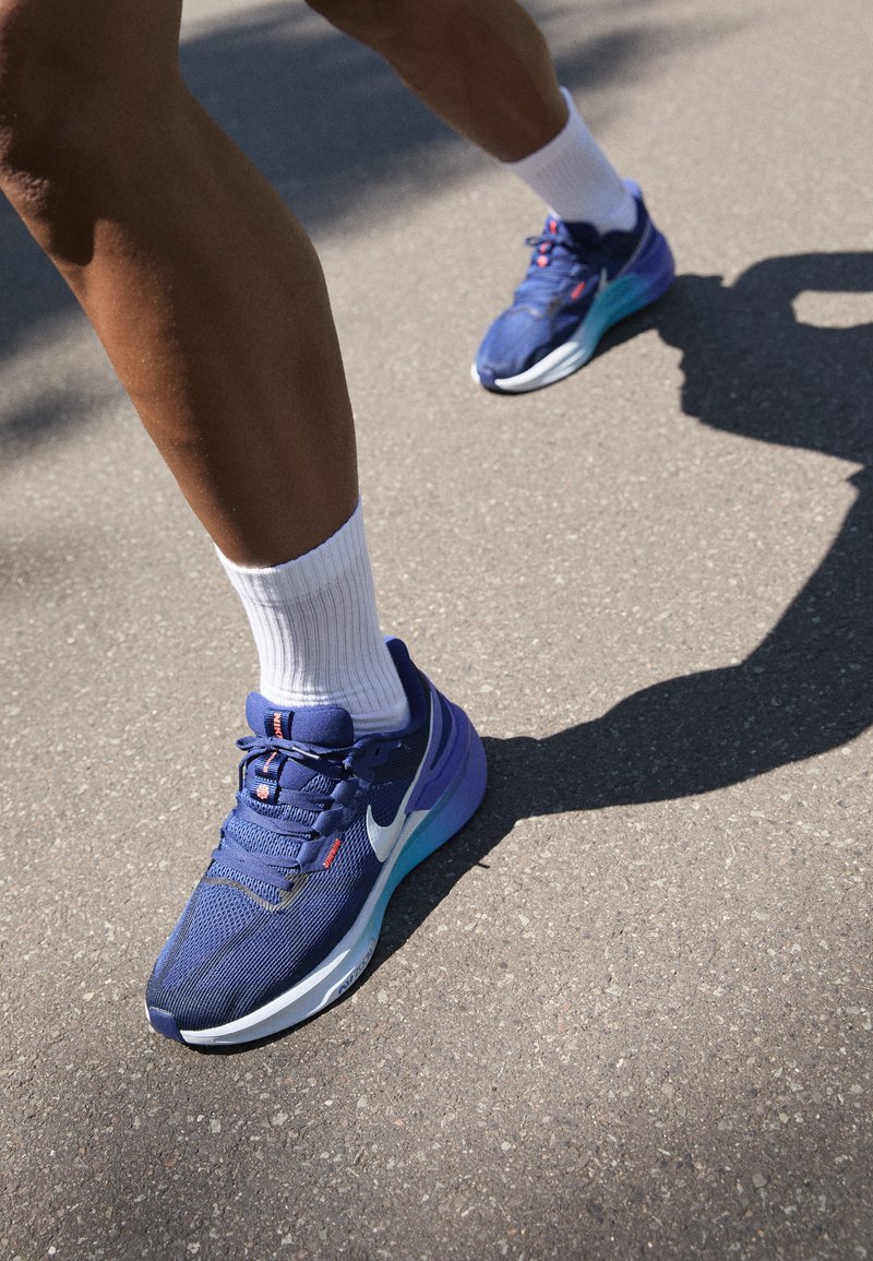 Nike tekaške čevlje v temno modri mrežasti zasnovi z belimi in vijoličnimi poudarki. Imajo oblazinjeno podplat in bele rebraste nogavice na asfaltni podlagi.