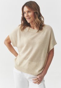 Maglione beige lavorato a maglia con maniche corte, scollo ampio e finitura testurizzata. Abbinato a pantaloni bianchi, il look è casual e rilassato.