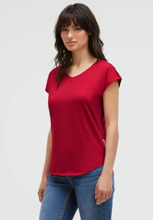 Woman with long dark hair wearing a red short-sleeve V-neck shirt and blue jeans standing against a plain light background.