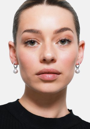 Silberne Creolen mit strukturiertem Design und einer einzelnen Perle, die am Boden baumelt. Das Modell trägt einen schwarzen Pullover.