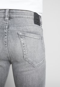 Šedé džíny z denimu s přiléhavým střihem, obsahující výrazné zadní kapsy a černou nášivku na pásku. Jemná textura a detaily prošití.