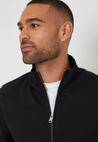 Zwarte zip-up jas met een hoge kraag en zilveren rits. Draagt over een witte t-shirt, die een gladde, textuurstof laat zien.