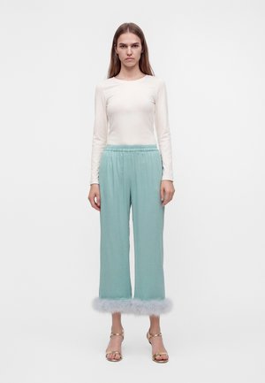 Le mannequin se tient devant un fond blanc, vêtue d’un ensemble deux pièces bleu aqua pâle orné de plumes couleur lavande et de sandales dorées à talons hauts.