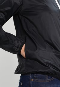 Černá lehká bunda s elastickými manžetami, předním zipem a bočními kapsami. Látka vypadá hladce a je voděodolná. Úzký střih.