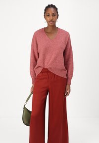 Roze wolblend trui met een V-hals en een iets losse pasvorm, gecombineerd met hoogpersoonlijke rode wijde broek. Accessoires omvatten een groene handtas.