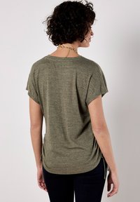 Camisa de color verde oliva, de manga corta, con un acabado texturizado, escote redondo y detalles fruncidos en los laterales para un toque de estilo. Combinada con jeans oscuros.