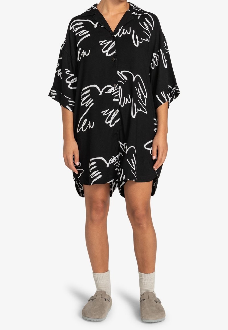 Personne portant une robe chemise noire boutonnée avec des motifs abstraits d'oiseaux blancs, associée à des chaussettes beiges et des chaussures sans lacets.