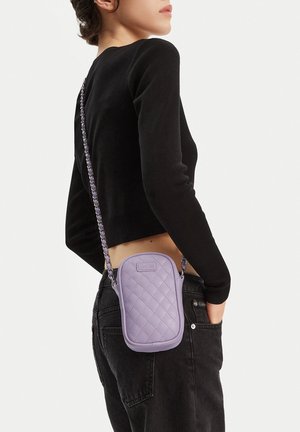 Kobieta ubrana w czarną bluzkę z długim rękawem i dżinsy, z małą pikowaną lawendową torebką na ramię przewieszoną od barku do biodra.