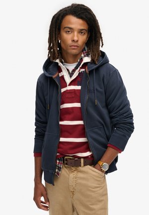 Marine blauwe zip-up hoodie met geribbelde manchetten, gedragen over een rood-wit gestreepte polo. Beige broek en een bruine riem maken de look compleet.