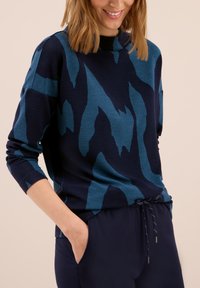 Pull bleu marine avec motif abstrait teal, texture côtelée, coupe décontractée et col montant avec détail de cordon au bas.