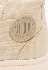 Baskets en toile beige dotées d'une semelle en caoutchouc texturée et d'une étiquette logo ronde et en relief avec l'inscription "PALLADIUM" et "DEPUIS 1947" embossée.
