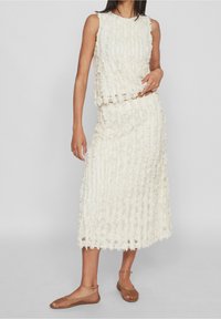 Kaksiosainen asu, joka koostuu hihattomasta topista ja midi-hameesta vaaleassa valkoisessa. Teksturoidusta kankaasta valmistettu, jossa on kukkakoristeita ja suora siluetti.