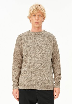 Junger Mann mit blondem Haar, der einen braun-weißen, strukturierten Strickpullover und schwarze Hosen trägt, steht vor einem schlichten hellen Hintergrund.