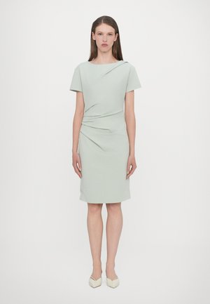 Mintgroene jurk tot de knie met korte mouwen en gedrapeerde details bij de taille. Gladde textuur, minimalistisch ontwerp, gecombineerd met lichtgekleurde hakken.
