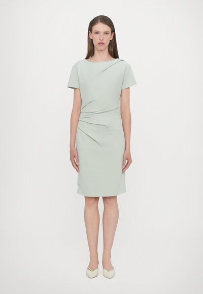 Robe midi vert menthe avec des manches courtes et des détails drapés à la taille. Texture lisse, design minimaliste, associée à des talons de couleur claire.