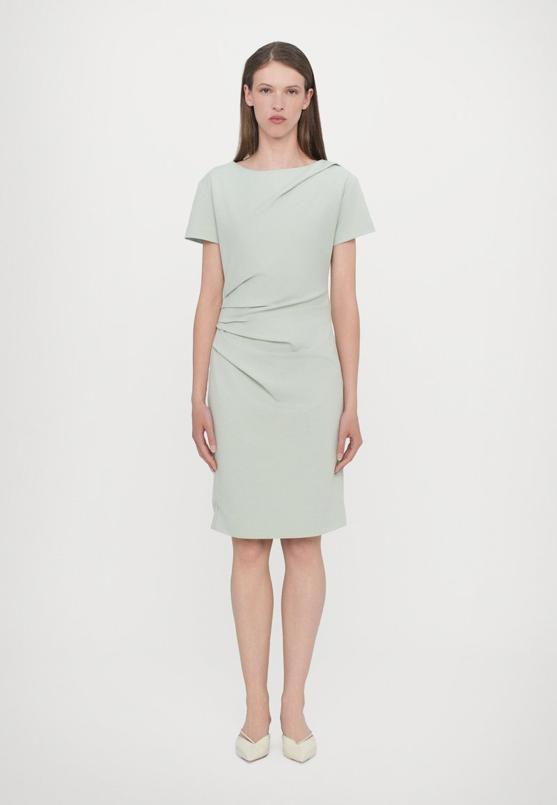 Rochie verde menta, până la genunchi, cu mâneci scurte și detalii drapate la talie. Textură netedă, design minimalist, asortată cu tocuri de culoare deschisă.