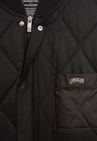 Svart quiltad jacka med dragkedja fram, ribbat krage och en ficka med ordet "ORÄTTVIS" i broderad skrift. Strukturerad tyg.