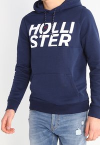 Sudadera azul marino con un gran logo blanco de "HOLLISTER", capucha con cordones, bolsillo tipo canguro en la parte frontal y puños y dobladillo acanalados.