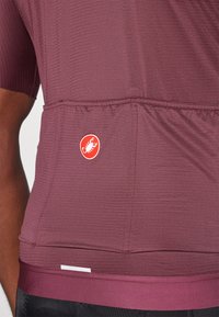 Burgundi sportpóló bordázott textúrával, rövid ujjakkal és testhez simuló fazonnal. Jellemzője egy logófelvarró és egy vízszintes varrás a stílus érdekében.
