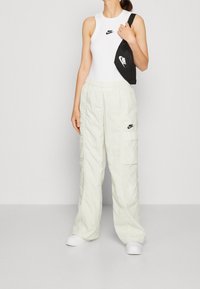 Vit ärmlös topp, ljusbeige cargobyxor och vita sneakers, med en svart axelväska som har logotypdetaljer på båda föremålen.