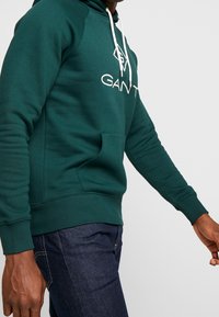 Sudadera verde de mezcla de algodón, con un bolsillo delantero, cordones blancos y un logotipo bordado en el pecho. Mangas y dobladillo con puños.