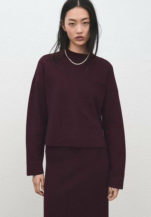 Ensemble pull bordeaux et jupe assortie ; matière en maille douce ; manches longues ; col rond ; texture subtile ; porté avec un collier chaîne argenté.